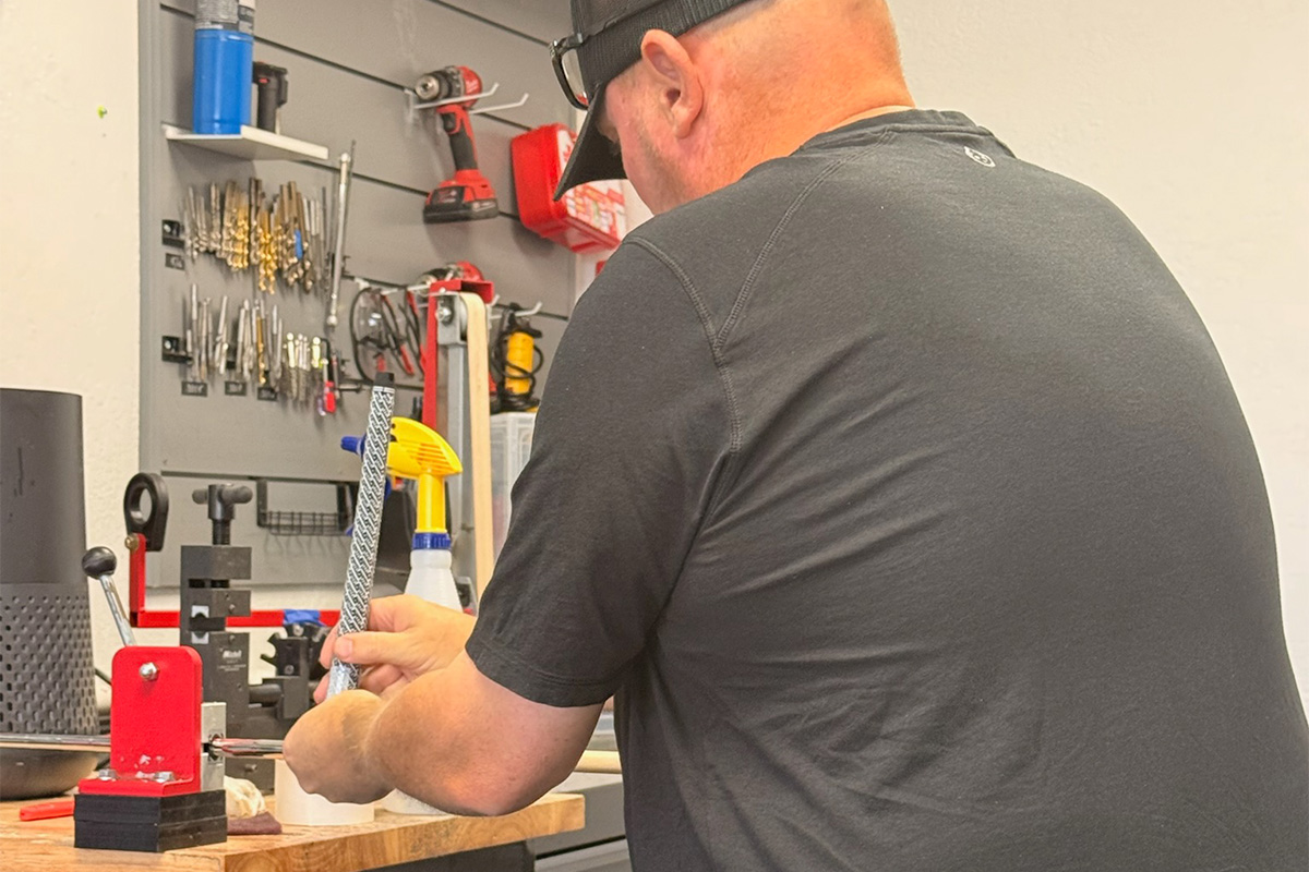 Man in a dark shirt and cap filing a metal rod clamped in a red bench vise at a cluttered workshop bench.