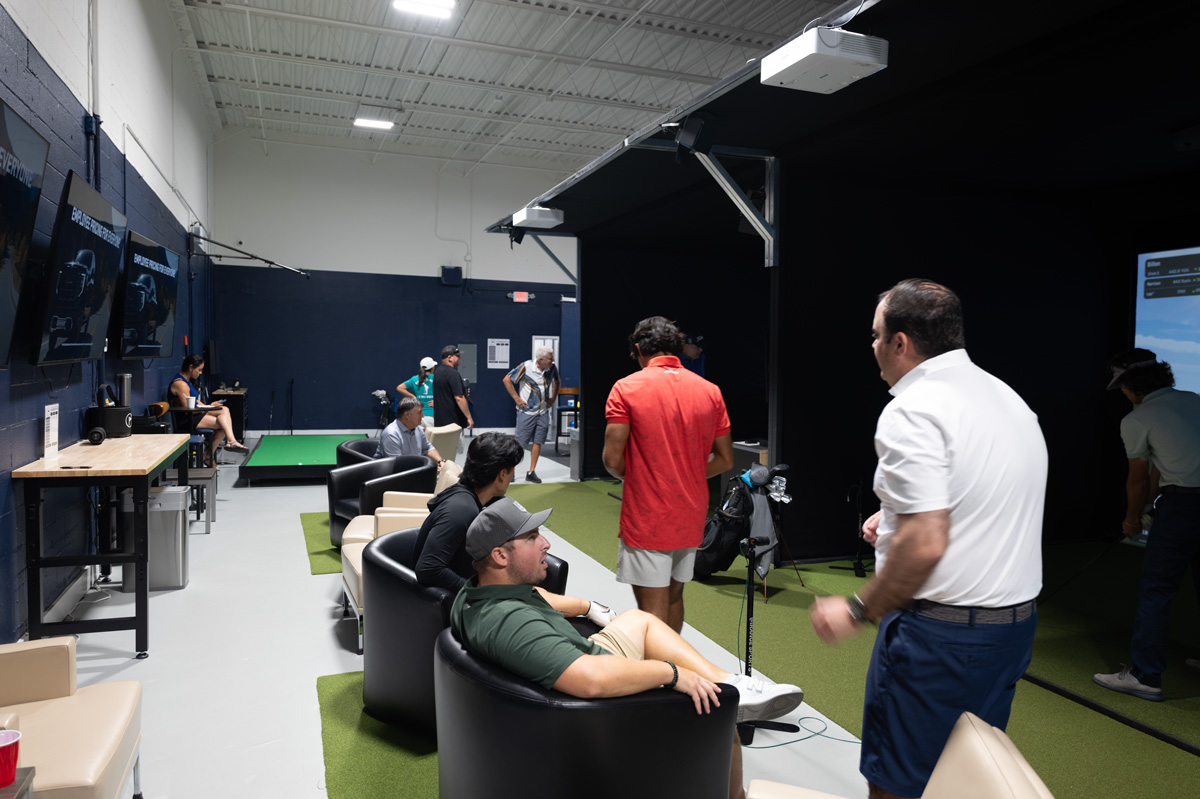 Indoor golf facility with people seated in lounge chairs while others stand near practice mats and simulators on the left wall-mounted screens fade into the background